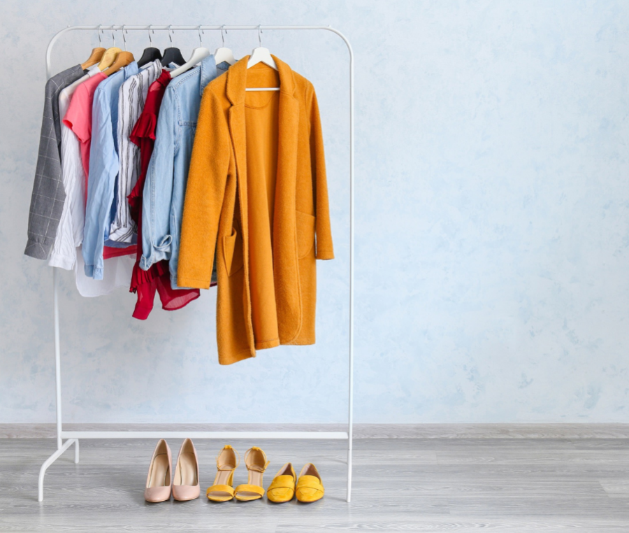 A white clothing rack holding a variety of colorful garments, with a prominent mustard yellow coat in the foreground. Below the rack, three pairs of yellow and beige shoes are arranged on a light gray wood floor against a pale blue textured wall.