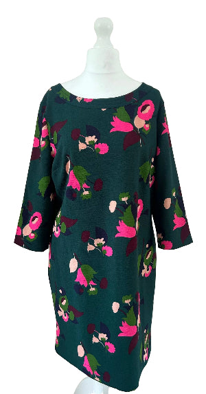 A dark green knee-length dress with three-quarter sleeves, featuring a vibrant pink, green, and purple floral pattern, displayed on a white mannequin against a plain beige wall.