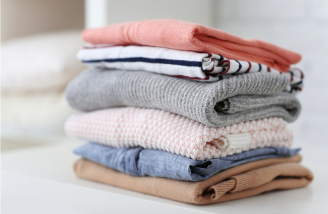 A stack of five folded knit sweaters in soft pastel colors, including coral pink, navy and white striped, heather gray, light pink, heather blue, and tan, arranged on a white surface.